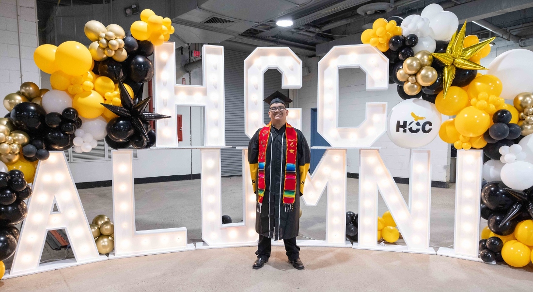 An HCC student poses for a photo in front of the HCC Alumni design at Fall Commencement.